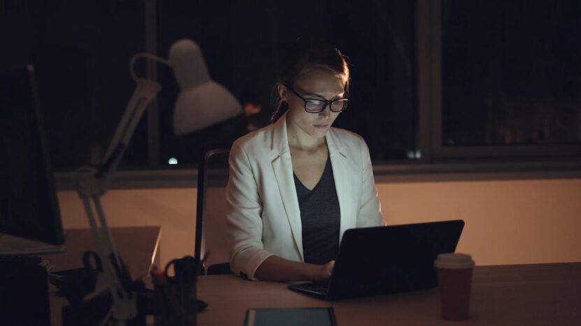 Woman wearing glasses working on a laptop at night in a dimly lit office