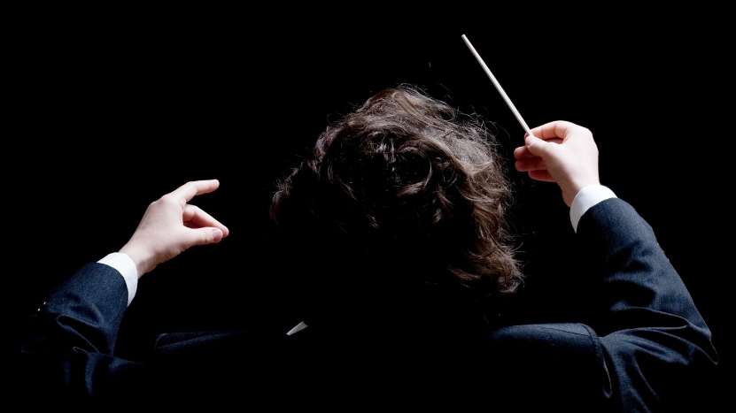 Orchestra conductor seen from behind, raising both hands with a baton to guide the performance in real time