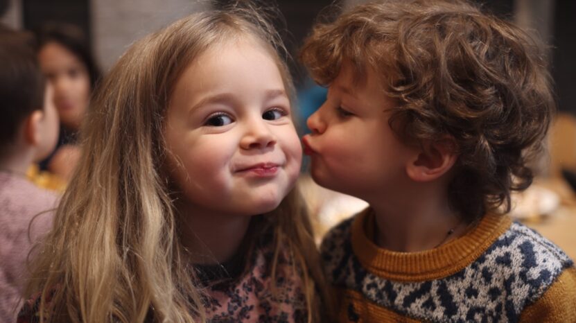 A young boy kissing a smiling girl on the cheek while sitting together, surrounded by other children in the background